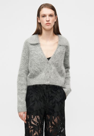 Γκρι, θολό πλεκτό ζακέτα με βαθύ V-λαιμό και γιακά, με κούμπωμα με κουμπιά. Συνδυάζεται με μαύρο παντελόνι με σχέδιο δαντέλας.