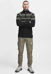 Schwarzer Strickpullover mit einem grünen und braunen geometrischen Muster, hohem Kragen, kombiniert mit olivgrünen Cargo-Hosen und grauen Sneakers.