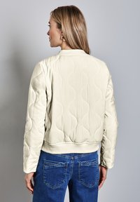 Beige gewatteerde bomberjack met een golvend patroon op de achterkant, geribbelde zoom en een ontspannen pasvorm. Gecombineerd met blauwe denim jeans.