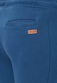 Blaue Sweatpants mit einer hinteren Tasche, die einen dezenten horizontalen Schlitz hat. Ein kleines braunes Lederetikett zeigt den Markennamen "FORSBERG".