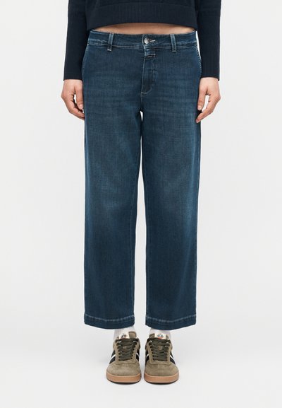 Person trägt dunkelblaue Jeans mit weitem Bein, schwarzes kurzes Oberteil, weiße Socken und beige Sneakers mit schwarzen Streifen.