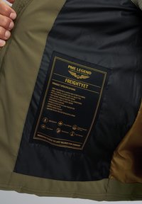 Green jacket with a black lining, featuring a care label with gold text detailing fabric composition and design specifications.