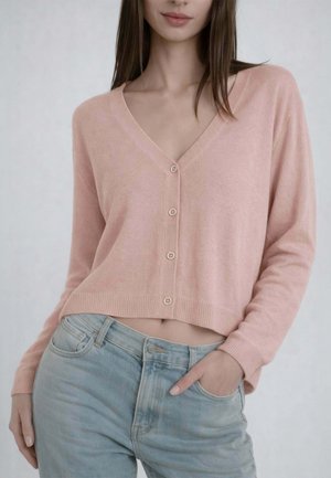Donna che indossa un cardigan rosa chiaro con bottoni e maniche lunghe, e jeans azzurri, con una mano nella tasca dei jeans, su uno sfondo semplice.