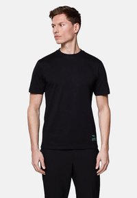 Czarna bawełniana koszulka z krótkimi rękawami i okrągłym dekoltem, zdobiona małym turkusowym logo u dołu, noszona z czarnymi spodniami.