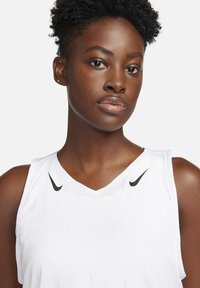 Haut de sport sans manches blanc avec un col en V ; possède de petits logos Nike noirs sur la poitrine, en tissu lisse et avec une texture minimale.