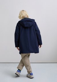 Persoon met kort blond haar, gekleed in een marineblauwe hoodiejas, beige broek en witte sneakers met blauwe accenten, die zijwaarts loopt op een lichte achtergrond.