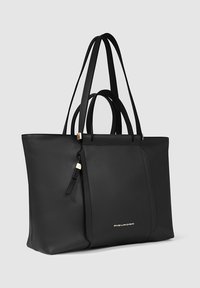 Bolso tote de cuero negro con doble asa, detalle de cremallera y logo dorado en el panel frontal, colocado contra un fondo neutro.