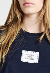 Una giovane persona con lunghi capelli biondi indossa una maglietta blu navy con una toppa bianca che riporta la scritta "Fidati del tempo della tua vita."