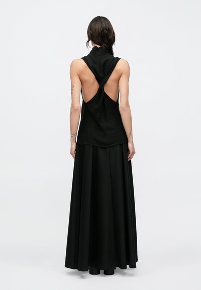 Černý top bez rukávů s vysokým límcem a otevřenými zády, s uzlovým detailem na zádech, spojený s dlouhou černou splývavou sukní.