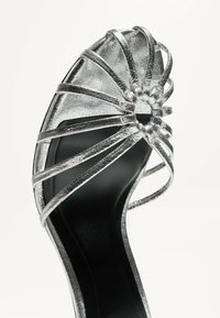 Silverfärgad metallisk högklackad sandal med flera tunna remmar och en cirkulär design vid tån, med en svart sula.