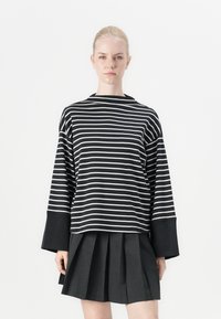 Blusa de mangas longas com riscas pretas e brancas, com gola alta e detalhes em preto nos punhos, combinada com uma saia plissada escura.