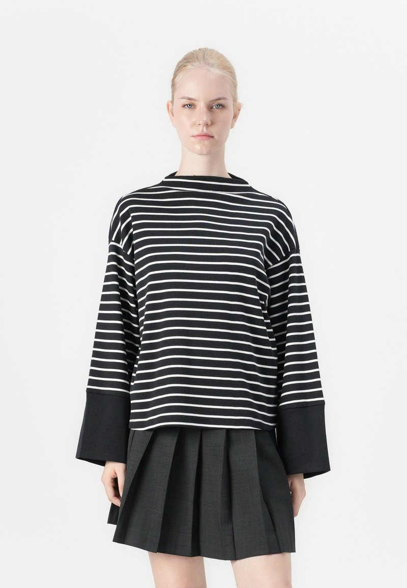 Blusa de mangas longas com riscas pretas e brancas, com gola alta e detalhes em preto nos punhos, combinada com uma saia plissada escura.