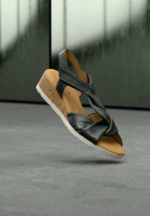 Μαύρο δερμάτινο σανδάλι με σχεδιασμό πλεγμένου λουριού, τακούνι από φελλό σε μορφή κύβου και λευκή σόλα πάνω σε γκρι επιφάνεια.