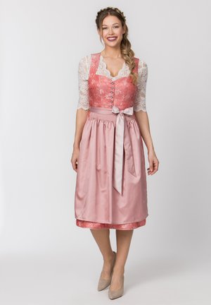 Frau lächelt, steht, trägt ein pinkfarbenes traditionelles Kleid mit Spitzärmeln, floralem Mieder, Satin-Schürze, die in der Taille gebunden ist, und beigefarbenen Absätzen.