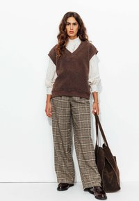 Pull en laine texturé marron sur une blouse à manches longues à motif écossais ; pantalon large à carreaux ; sac marron foncé ; chaussures noires brillantes.