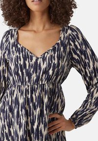 Robe avec un motif abstrait bleu marine et crème, décolleté en V, et manches longues avec des poignets froncés, fabriquée en tissu léger.