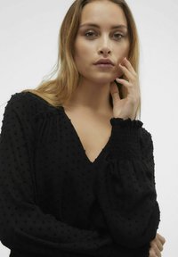 Blusa nera con texture, caratterizzata da scollo a V, maniche a sbuffo con polsini elasticizzati e piccole pallini in rilievo su tutta la stoffa.