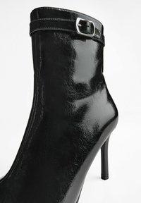 Ghetă de gleznă din piele lacuită neagră, cu un design elegant, toc înalt și un accesoriu cu cataramă pe manșetă. Finisaj texturat și lucios.