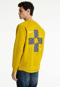 Sudadera amarilla con cuello redondo, que presenta un diseño geométrico en blanco y negro en la parte posterior. Textura suave, corte informal, puños de canalé.