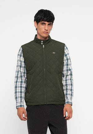 Chaleco acolchado verde con cuello alto y cierre frontal. Lleva puesto una camisa de cuadros azul y blanca. Presenta un pequeño logo en el lado izquierdo del pecho.