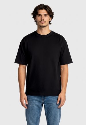 Giovane uomo con capelli scuri di media lunghezza, che indossa una semplice t-shirt nera a maniche corte e jeans blu, in piedi davanti a uno sfondo chiaro.