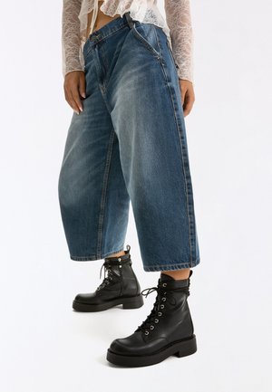 Personne portant un jean bleu à jambes larges et coupé, des bottines noires à lacets et un haut blanc en dentelle à manches longues avec un ourlet volanté.
