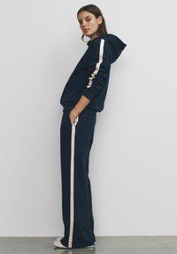Woman standing sideways wearing a navy blue tracksuit with beige stripes down the sleeves and pant legs, hands in pockets, against a plain wall.