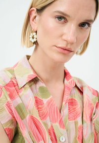 Camicia colorata e decorata con onde rosa e verdi. Maniche corte e colletto, con un dettaglio di bottone bianco. Orecchini simili a perle indossati.