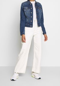 Jeansjacka i mörkblått med silverknappar och två framfickor, kombinerad med beige vida byxor och vita sneakers.