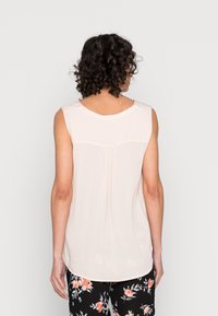 Blusa sin mangas de color rosa claro con escote redondo, textura suave y detalle de plisado en la parte posterior, combinada con pantalones negros de estampado floral.