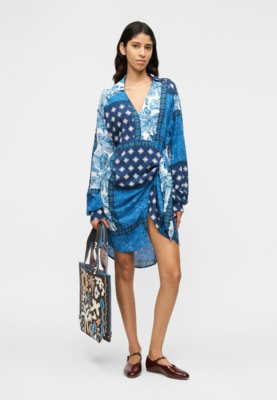 Μπλε φόρεμα με σχέδια, μακριά μανίκια, V-λαιμόκοψη και ζώνη στη μέση. Το μοντέλο κρατά καφέ τσάντα ώμου με πολύχρωμα γραφικά σχέδια.
