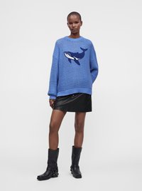 Personne portant un pull en maille bleu avec un motif de baleine, une mini-jupe en cuir noire et des bottes noires mi-mollet, debout devant un fond uni.