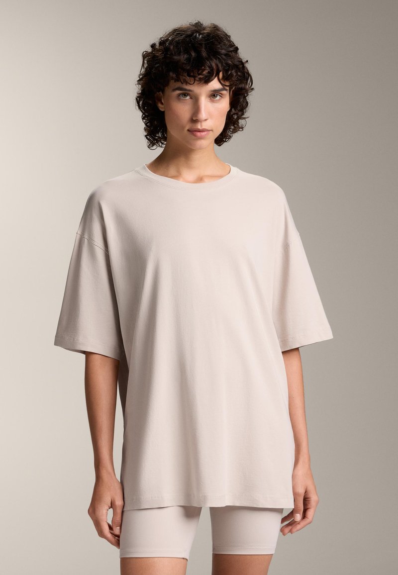 Bež majica oversized kroja izrađena od mekog materijala, sa okruglim izrezom i kratkim rukavima, u kombinaciji sa svijetlim šortsevima.