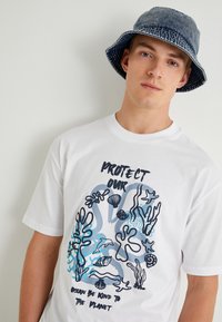 Camiseta blanca de algodón con un diseño gráfico audaz que presenta ilustraciones de vida marina en tonos de azul y negro. El texto dice: "Protege nuestro océano, sé amable con el planeta."