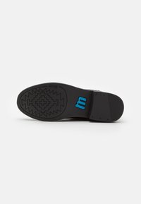 Suela de zapato negra con un dibujo de zigzag, con un logo azul "m" y acentos texturizados. El diseño incluye áreas lisas y estriadas para mayor agarre.