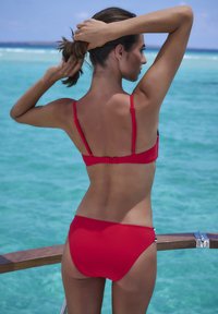Rdeč bikini zgoraj z tankimi naramnicami in zapenjanjem na hrbtu, v kombinaciji z ustreznimi hlačkami z visokim pasom. Gladek, elastičen material ob koži.