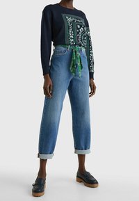 Marineblauwe sweatshirt met groene patronen, gecombineerd met lichtblauwe, hooglopende spijkerbroek. Blauwe loafers met een dikke zool maken de look compleet.