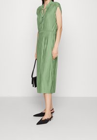 Grünes, plissiertes Midikleid mit kurzen Ärmeln, Schlüsselloch-Ausschnitt, tailliertem Bund und schwarzen Slingback-Schuhen mit spitzem Fuß.