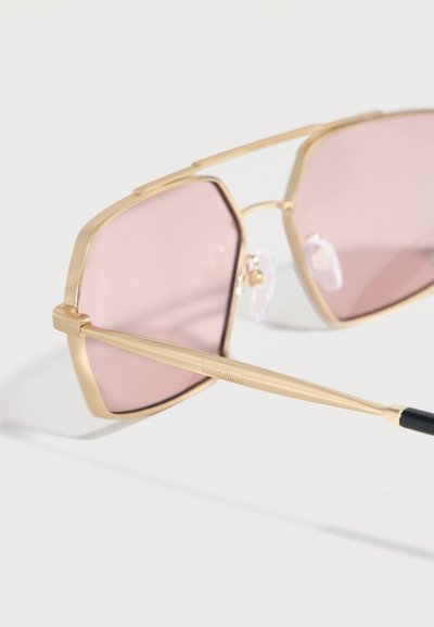 Γυαλιά ηλίου με γεωμετρικά πλαίσια σε χρυσό τόνο και φακούς με ροζ απόχρωση, τοποθετημένα πάνω σε λευκή επιφάνεια.