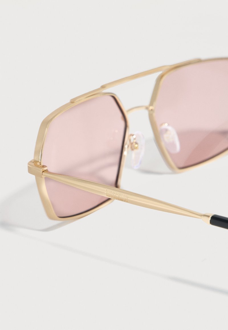 Gafas de sol con montura geométrica dorada y lentes tintados en rosa, descansando sobre una superficie blanca.