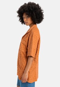 Short-sleeve, burnt orange shirt with a textured floral pattern, featuring a chest pocket and a relaxed fit design.