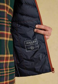 Námořnická modrá podšívka s oranžovým lemem a štítkem Joules. Kostičkovaná vnější tkanina v zelené, žluté a červené barvě. Texturovaný povrch, zipové uzavření.