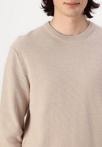 Jong persoon in een beige gebreide trui met een ronde hals tegen een effen witte achtergrond.