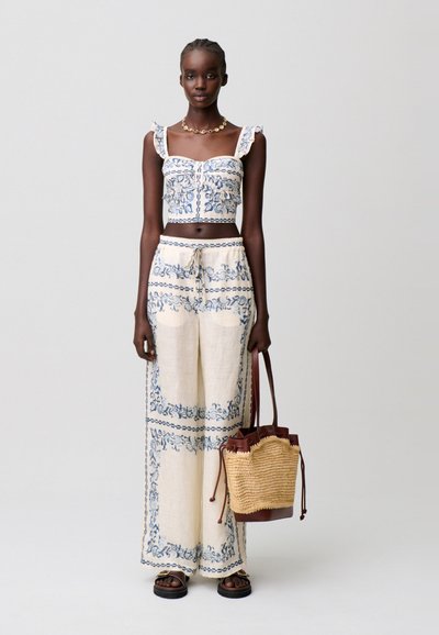 Donna che indossa un completo crema a due pezzi con motivi floreali blu, tiene una borsa intrecciata con dettagli in pelle, in piedi davanti a uno sfondo semplice.
