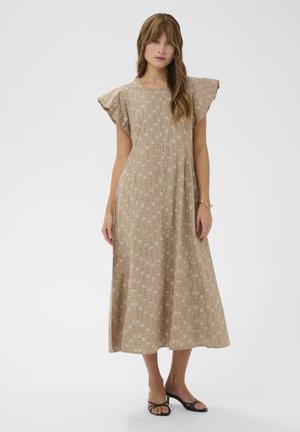 Kobieta stojąca w beżowej sukience midi z falbanowymi rękawami, pionowym małym kwiatowym wzorem, czarnych butach na obcasie z odkrytymi palcami oraz złotej bransoletce na nadgarstku.
