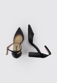 Zapatos de tacón altos negros con punta afilada y tira en el tobillo. Cuentan con un tacón grueso y una textura de cuero suave. Suela interior de color claro visible.