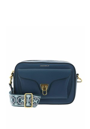 Sac bandoulière en cuir bleu marine avec poche à rabat sur le devant, fermoir doré et sangle réglable en tissu à motifs. Nom de la marque "Coccinelle" inscrit en haut.