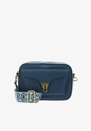 Sac bandoulière en cuir bleu marine avec poche à rabat sur le devant, fermoir doré et sangle réglable en tissu à motifs. Nom de la marque "Coccinelle" inscrit en haut.