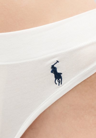 Κοντινή λήψη της μέσης λευκού εσωρούχου με σκούρο μπλε κεντημένο λογότυπο παίκτη πόλο σε φόντο ανοιχτόχρωμης επιδερμίδας.