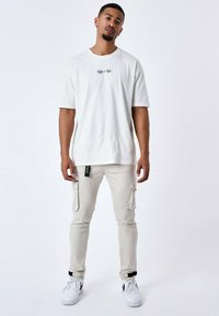 Camiseta blanca de algodón con logo negro; pantalones cargo color beige claro con bolsillos; etiqueta negra en el costado; zapatillas deportivas blancas con detalles negros.